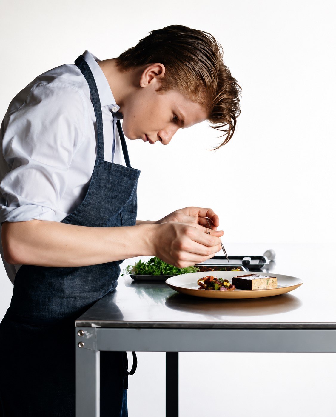 A young chef plating a dish on a stainless-steel worktop