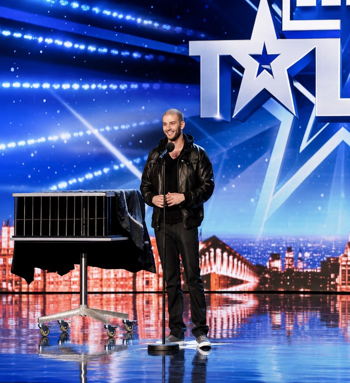 A young magician at the microphone on a talent-show stage, a covered cage beside him