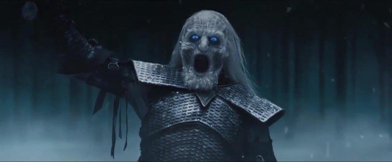A pale, blue-eyed warrior in dark armour standing in a dark snowy wood, mouth open in a scream, one arm raised holding a sword and the other held out from his body.